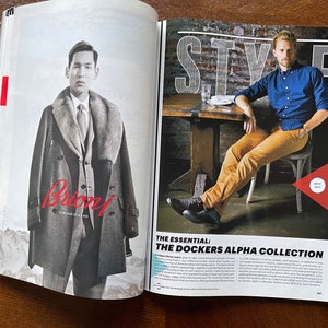 May include: A magazine spread featuring two men in different styles. The man on the left is wearing a gray coat with a fur collar and the text "Brioni" in red. The man on the right is wearing a blue button-down shirt, khaki pants, and black shoes. The text "THE ESSENTIAL: THE DOCKERS ALPHA COLLECTION" is visible in the bottom right corner.