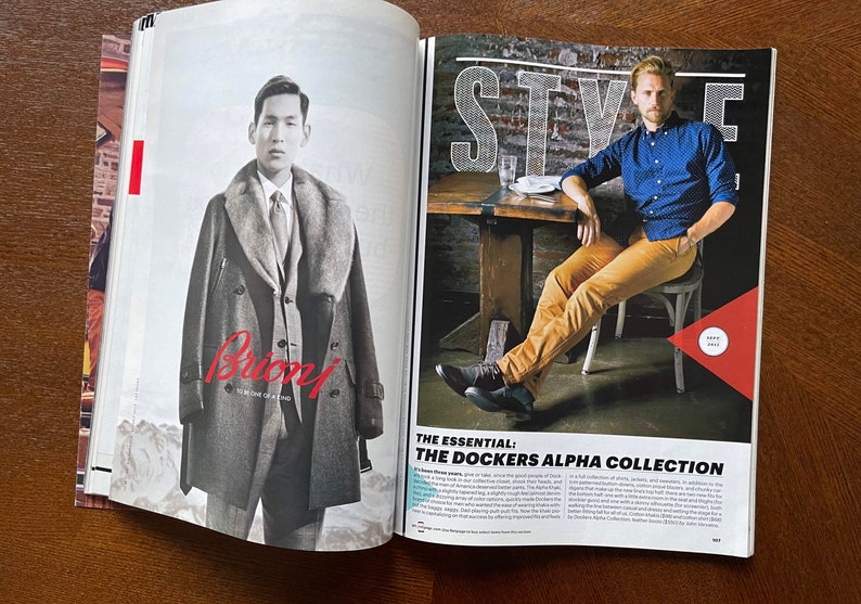 May include: A magazine spread featuring two men in different styles. The man on the left is wearing a gray coat with a fur collar and the text "Brioni" in red. The man on the right is wearing a blue button-down shirt, khaki pants, and black shoes. The text "THE ESSENTIAL: THE DOCKERS ALPHA COLLECTION" is visible in the bottom right corner.