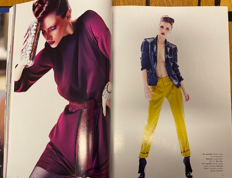 May include: Two women modelling fashion. The woman on the left is wearing a burgundy dress with a gold belt and a silver clutch. The woman on the right is wearing a black sequined jacket, yellow trousers, and black heels.
