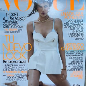 Op de afbeelding: Een Vogue España tijdschriftomslag met een persoon die een wit strapless topje met dunne bandjes, een gouden ketting en een bloemenhoofdtooi draagt. De omslag bevat dikke oranje tekst, waaronder de titel VOGUE en diverse Spaanstalige koppen.