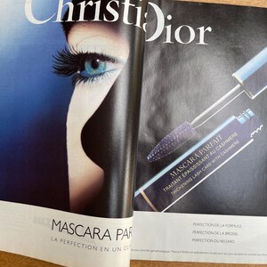 May include: An open magazine featuring a Christian Dior mascara advertisement. The ad showcases a close-up of a woman's eye with long, dark lashes and a blue eye shadow. The mascara tube and applicator are also visible.