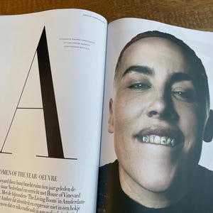 May include: A magazine spread featuring a portrait of a person with a shaved head and a black top. The person is smiling, revealing silver teeth. The left page has a large black "A" and text about "WOMEN OF THE YEAR OEUVRE".