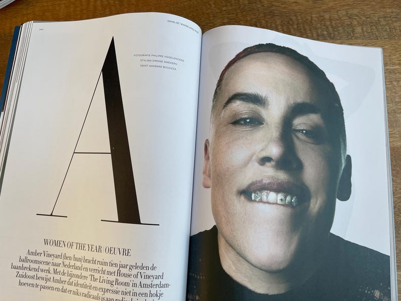 May include: A magazine spread featuring a portrait of a person with a shaved head and a black top. The person is smiling, revealing silver teeth. The left page has a large black "A" and text about "WOMEN OF THE YEAR OEUVRE".