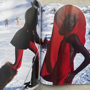 Puede incluir: Una revista de moda con una modelo con un vestido rojo y una gran capucha roja. La modelo est&aacute; de pie frente a un fondo de monta&ntilde;a nevada. Otra imagen muestra a un esquiador con atuendo negro y rojo con botas forradas de piel y esqu&iacute;s.