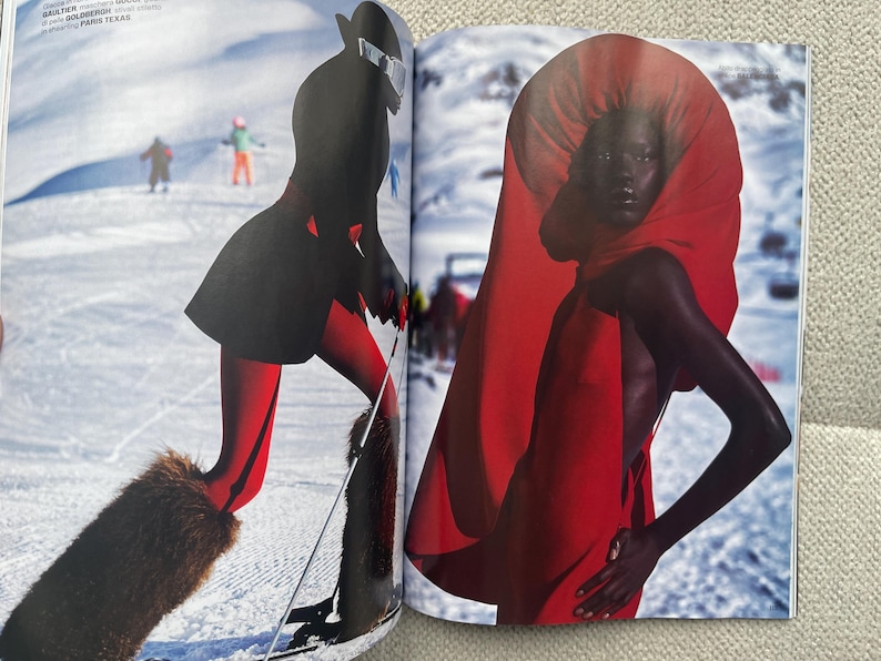 Puede incluir: Una revista de moda con una modelo con un vestido rojo y una gran capucha roja. La modelo est&aacute; de pie frente a un fondo de monta&ntilde;a nevada. Otra imagen muestra a un esquiador con atuendo negro y rojo con botas forradas de piel y esqu&iacute;s.