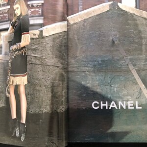 以下が含まれることがあります： フリンジトリムと茶色のベルトが付いた黒いドレスを着た女性が屋上に立っています。背景はレンガの壁と、「CHANEL」という文字が印刷されたコンクリートの壁です。