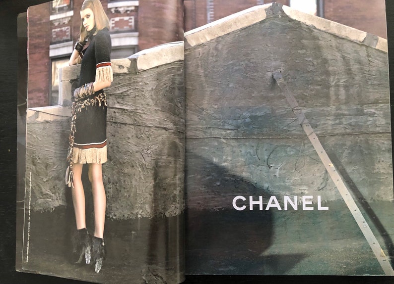 以下が含まれることがあります： フリンジトリムと茶色のベルトが付いた黒いドレスを着た女性が屋上に立っています。背景はレンガの壁と、「CHANEL」という文字が印刷されたコンクリートの壁です。