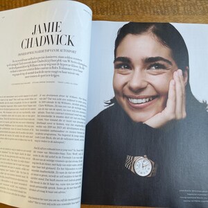 May include: Magazine spread featuring a smiling woman wearing a black turtleneck and a gold wristwatch. The watch has a white face and a gold band. The magazine's headline reads "Jamie Chadwick".