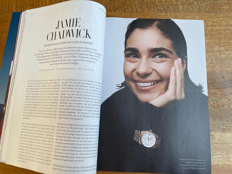May include: Magazine spread featuring a smiling woman wearing a black turtleneck and a gold wristwatch. The watch has a white face and a gold band. The magazine's headline reads "Jamie Chadwick".