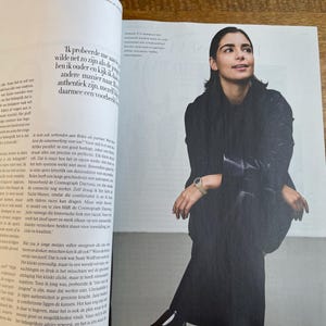 May include: A woman in a black outfit, seated and looking upwards, is featured in a magazine spread. She wears a watch with a gold band. The image is part of a fashion or lifestyle editorial.