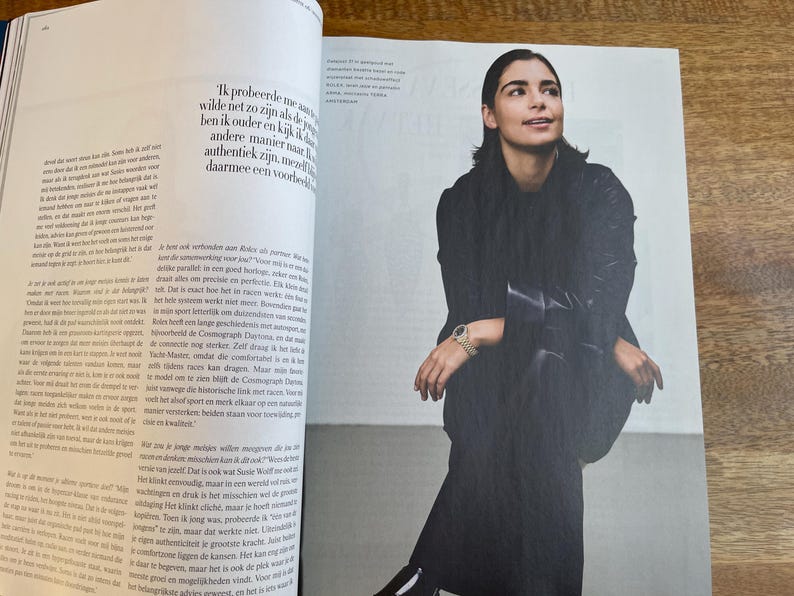 May include: A woman in a black outfit, seated and looking upwards, is featured in a magazine spread. She wears a watch with a gold band. The image is part of a fashion or lifestyle editorial.