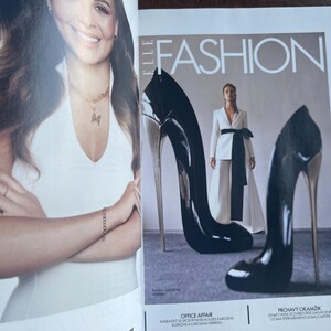 May include: A magazine cover featuring the word "FASHION" in white capital letters. Two oversized black high heels with silver heels are in the foreground. A woman in a white suit with a black sash is in the background.