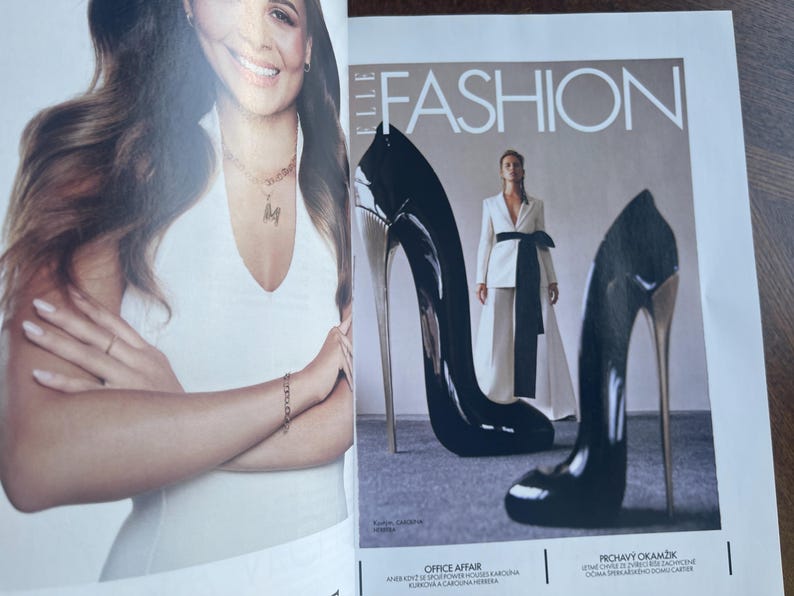 May include: A magazine cover featuring the word "FASHION" in white capital letters. Two oversized black high heels with silver heels are in the foreground. A woman in a white suit with a black sash is in the background.