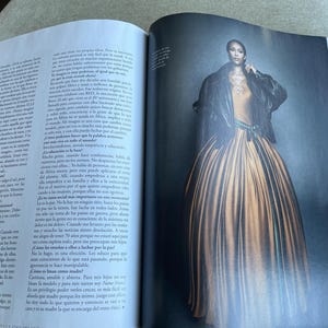Puede incluir: Una p&aacute;gina de revista de moda con una modelo con un vestido largo y fluido de color amarillo dorado. La modelo lleva una chaqueta negra sobre una parte superior de color claro. El vestido tiene una falda ancha y est&aacute; ce&ntilde;ido a la cintura con una faja verde. El fondo es oscuro.