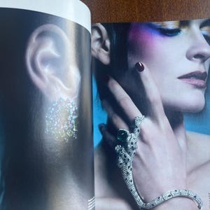 May include: A fashion magazine spread featuring a woman with dramatic makeup and jewelry. A silver bracelet and ring set with emerald and clear stones adorn the hand. The ear on the left side of the image has a sparkling earring.
