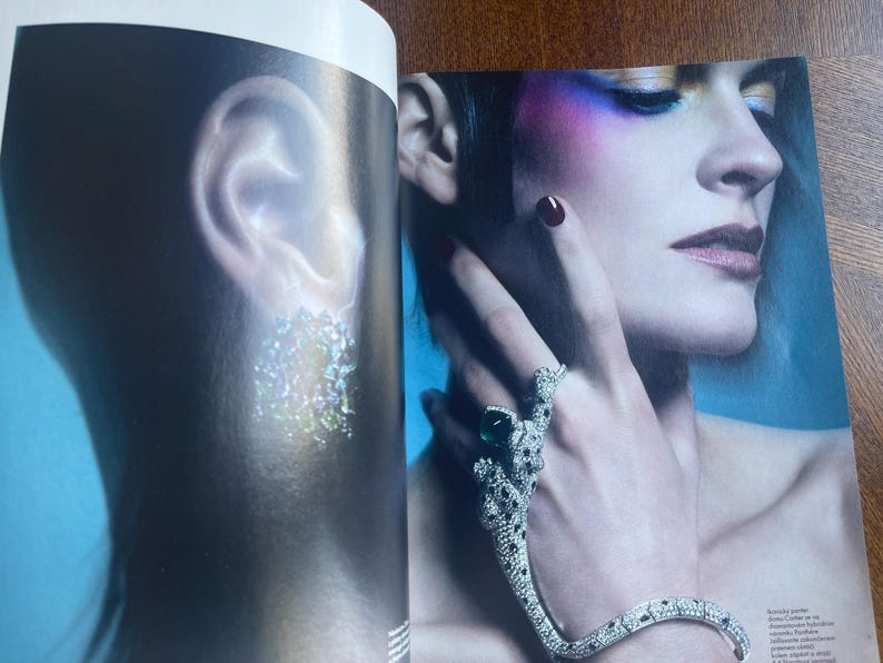 May include: A fashion magazine spread featuring a woman with dramatic makeup and jewelry. A silver bracelet and ring set with emerald and clear stones adorn the hand. The ear on the left side of the image has a sparkling earring.