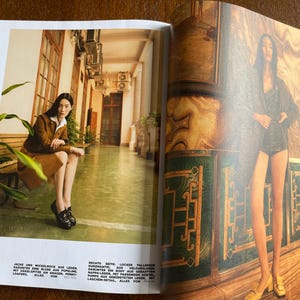 Puede incluir: Una revista de moda de doble p&aacute;gina. Una mujer con chaqueta y falda de ante marr&oacute;n, sentada en un banco. La otra p&aacute;gina muestra a una mujer con chaqueta y pantalones cortos oscuros. El fondo incluye detalles arquitect&oacute;nicos y texto.