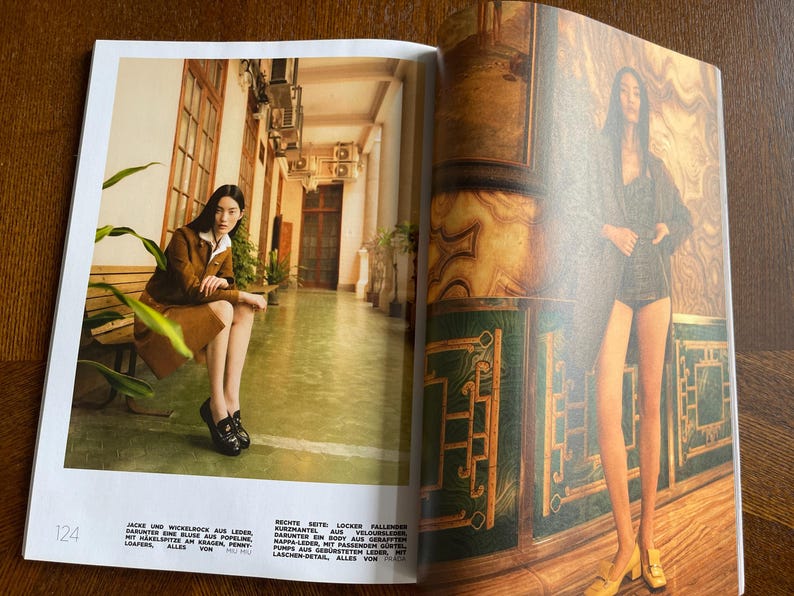 Puede incluir: Una revista de moda de doble p&aacute;gina. Una mujer con chaqueta y falda de ante marr&oacute;n, sentada en un banco. La otra p&aacute;gina muestra a una mujer con chaqueta y pantalones cortos oscuros. El fondo incluye detalles arquitect&oacute;nicos y texto.