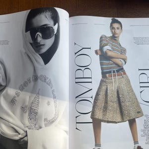 May include: A magazine spread with a model in sunglasses and a hoodie with the text "IT'S NOT ROCK N ROLL". The other model wears a striped shirt, pleated skirt, and the text "TOMBOY GIRL".