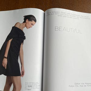 Puede incluir: Una p&aacute;gina de revista de moda con una mujer con un vestido negro y capa. La palabra "BEAUTIFUL" se muestra en grande. La revista est&aacute; abierta sobre una superficie de madera.