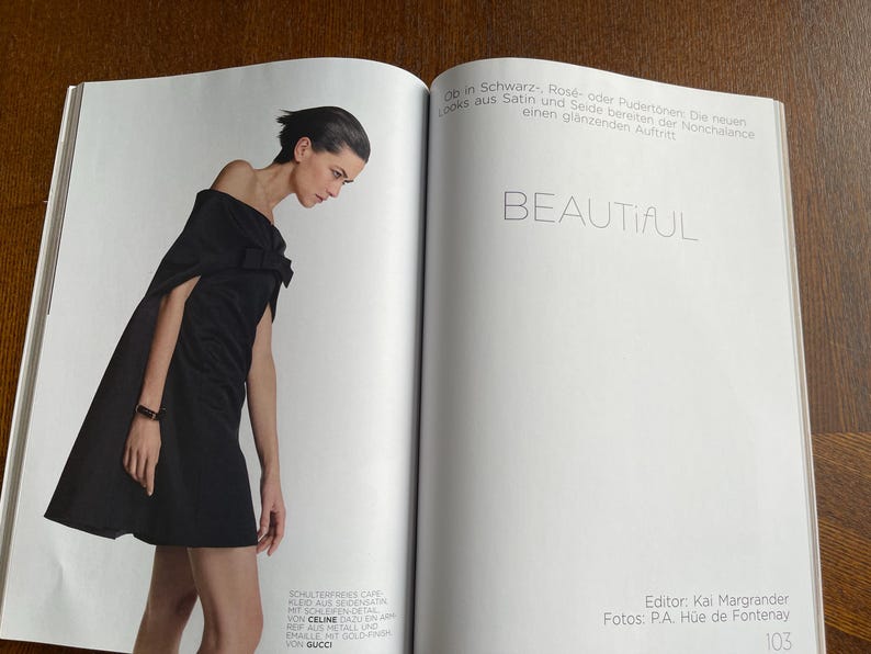 Puede incluir: Una p&aacute;gina de revista de moda con una mujer con un vestido negro y capa. La palabra "BEAUTIFUL" se muestra en grande. La revista est&aacute; abierta sobre una superficie de madera.