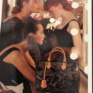 以下が含まれることがあります： 黒いドレスを着た3人の女性が、電球のついた鏡の前に立っています。手前にいる女性は黒い手袋をして、花柄の黒と茶色のルイ・ヴィトンのハンドバッグを持っています。