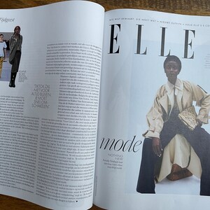 Puede incluir: Una portada de revista con la palabra "ELLE" en letras grandes y en negrita. La portada presenta a una modelo con un abrigo beige y pantalones oscuros, sentada en un taburete blanco. La modelo sostiene un peque&ntilde;o embrague estampado. El t&iacute;tulo de la revista es "mode" y el texto "NOTHING NEW" est&aacute; debajo.