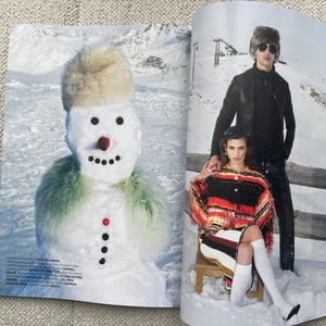 Puede incluir: Una p&aacute;gina de revista de moda con un mu&ntilde;eco de nieve y dos personas con ropa de invierno. El mu&ntilde;eco de nieve tiene un sombrero de piel, una nariz roja y una bufanda verde. Las personas llevan abrigos, gafas de sol y botas en un paisaje nevado.