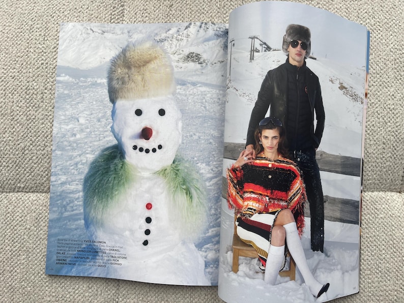 Puede incluir: Una p&aacute;gina de revista de moda con un mu&ntilde;eco de nieve y dos personas con ropa de invierno. El mu&ntilde;eco de nieve tiene un sombrero de piel, una nariz roja y una bufanda verde. Las personas llevan abrigos, gafas de sol y botas en un paisaje nevado.