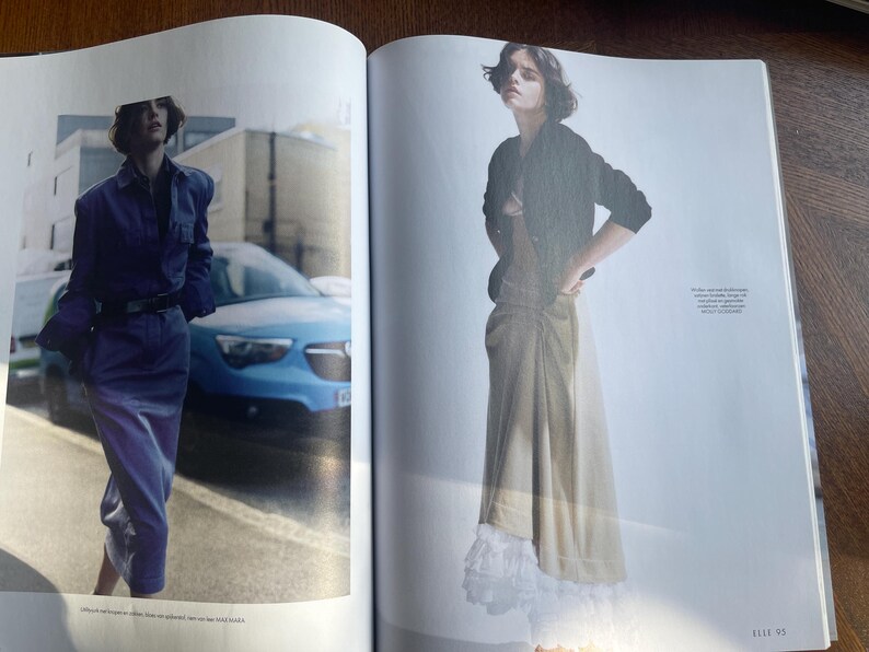 May include: A double-page spread from a fashion magazine. One model is in a blue denim ensemble, the other in a black jacket and a long beige skirt. A blue car and a white wall are visible in the background.