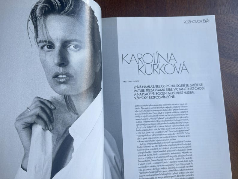 May include: Black and white magazine spread featuring a portrait of Karolina Kurkova. She is wearing a white collared shirt and a ring. The magazine page includes the text "ROZHOVOR Elle" and "KAROLINA KURKOV&Aacute;".