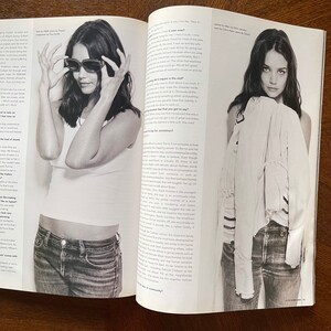 May include: A black and white photo of a woman wearing a white t-shirt and blue jeans. She is looking at the camera and holding her sunglasses in her hands. The photo is from a magazine and includes text about the woman.