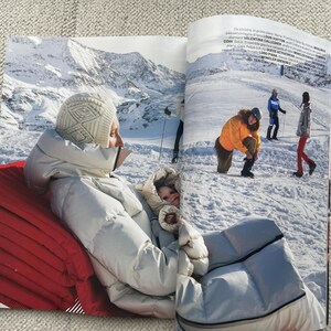 Puede incluir: Una p&aacute;gina de revista con moda de invierno. Una persona con una chaqueta acolchada y gorro blancos est&aacute; sentada en un coj&iacute;n rojo, con un fondo de monta&ntilde;as nevadas. Otras personas est&aacute;n en una pista de esqu&iacute;, vestidas con ropa de invierno.