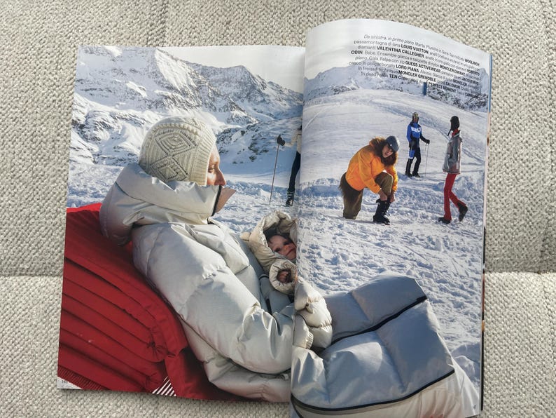 Puede incluir: Una p&aacute;gina de revista con moda de invierno. Una persona con una chaqueta acolchada y gorro blancos est&aacute; sentada en un coj&iacute;n rojo, con un fondo de monta&ntilde;as nevadas. Otras personas est&aacute;n en una pista de esqu&iacute;, vestidas con ropa de invierno.