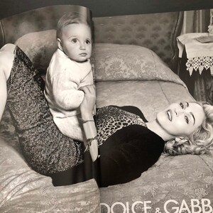 以下が含まれることがあります： ベッドに横たわる女性と、その膝の上に座っている幼い子供の白黒写真。女性はヒョウ柄のトップスと黒いスカートを着用している。子供は白いセーターを着ている。写真は、ナイトスタンドとランプが置かれた寝室で撮影された。