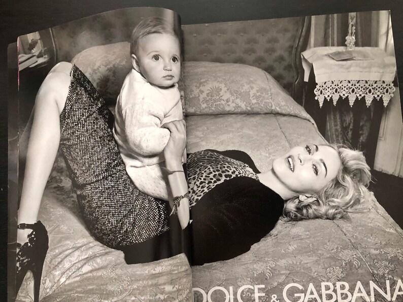 以下が含まれることがあります： ベッドに横たわる女性と、その膝の上に座っている幼い子供の白黒写真。女性はヒョウ柄のトップスと黒いスカートを着用している。子供は白いセーターを着ている。写真は、ナイトスタンドとランプが置かれた寝室で撮影された。