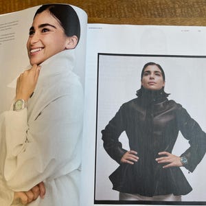 May include: Two-page spread from a magazine featuring a woman wearing a white coat and a black leather jacket. The woman is wearing a watch on her wrist in both images. The magazine pages are on a wooden surface.