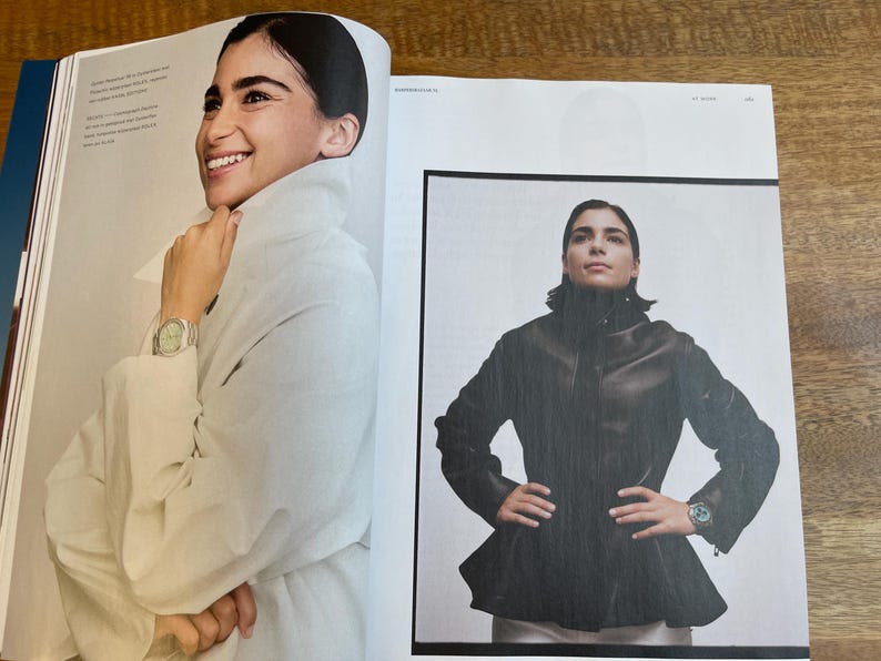 May include: Two-page spread from a magazine featuring a woman wearing a white coat and a black leather jacket. The woman is wearing a watch on her wrist in both images. The magazine pages are on a wooden surface.