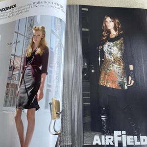 May include: Two women modeling fashion apparel in a magazine spread. One woman wears a dark burgundy cardigan, a black dress, and a brown belt. The other woman wears a patterned tunic, black leggings, and knee-high boots. The word "AIRFIELD" is visible.