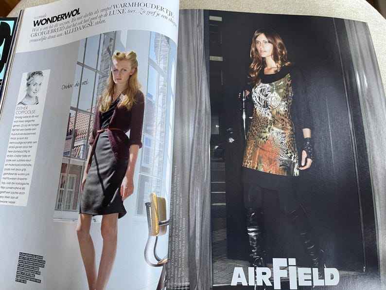 May include: Two women modeling fashion apparel in a magazine spread. One woman wears a dark burgundy cardigan, a black dress, and a brown belt. The other woman wears a patterned tunic, black leggings, and knee-high boots. The word "AIRFIELD" is visible.