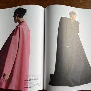 Puede incluir: Una revista de moda de doble p&aacute;gina. Una modelo lleva un abrigo &oacute;pera largo y rosa, la otra un abrigo con capa gris oscuro. El texto en la p&aacute;gina izquierda dice "N&deg;21 BY ALESSANDRO DELL'ACQUA" y "BALENCIAGA".