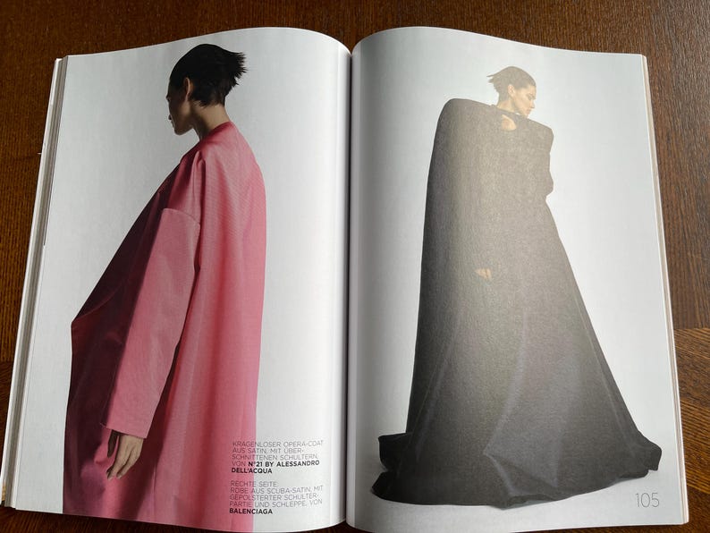 Puede incluir: Una revista de moda de doble p&aacute;gina. Una modelo lleva un abrigo &oacute;pera largo y rosa, la otra un abrigo con capa gris oscuro. El texto en la p&aacute;gina izquierda dice "N&deg;21 BY ALESSANDRO DELL'ACQUA" y "BALENCIAGA".