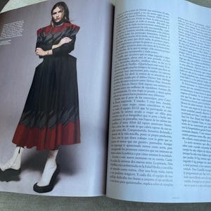 Puede incluir: Una imagen editorial de moda con una modelo con un vestido negro y rojo con un dise&ntilde;o &uacute;nico. El vestido tiene un cuello alto y una falda acampanada. La modelo lleva botas blancas con tacones negros. La imagen es de una revista.