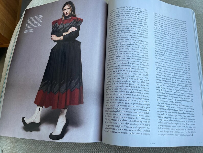 Puede incluir: Una imagen editorial de moda con una modelo con un vestido negro y rojo con un dise&ntilde;o &uacute;nico. El vestido tiene un cuello alto y una falda acampanada. La modelo lleva botas blancas con tacones negros. La imagen es de una revista.