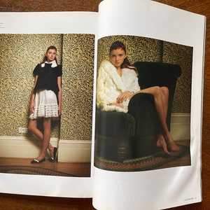May include: Two young women wearing different outfits pose for a photo shoot. One woman is wearing a white skirt with a black top and black shoes. The other woman is wearing a white fur coat and sitting on a dark green armchair. Both women are in a room with a patterned wallpaper.