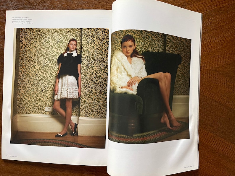 May include: Two young women wearing different outfits pose for a photo shoot. One woman is wearing a white skirt with a black top and black shoes. The other woman is wearing a white fur coat and sitting on a dark green armchair. Both women are in a room with a patterned wallpaper.