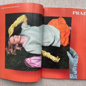 Puede incluir: Una p&aacute;gina de revista de moda con una mujer con un atuendo verde claro y guantes amarillos, acostada sobre la hierba. Tambi&eacute;n se ve una prenda naranja y una bolsa rosa. La palabra "PRADA" est&aacute; impresa en blanco.