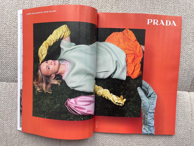 Puede incluir: Una p&aacute;gina de revista de moda con una mujer con un atuendo verde claro y guantes amarillos, acostada sobre la hierba. Tambi&eacute;n se ve una prenda naranja y una bolsa rosa. La palabra "PRADA" est&aacute; impresa en blanco.