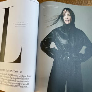 May include: A magazine spread featuring a woman in a black leather coat with a belt. The left page has a large black letter "L" and text that reads "WOMEN OF THE YEAR INTERNATIONAAL". The woman is smiling.