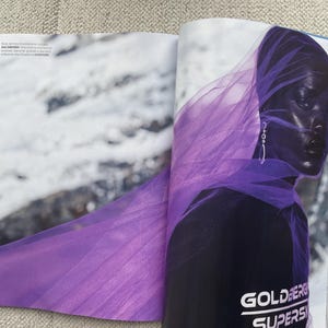 Puede incluir: Una p&aacute;gina de revista de moda con una persona de piel oscura, envuelta en una tela p&uacute;rpura transparente y fluida. La persona lleva pendientes plateados. El t&iacute;tulo de la revista, "GOLDBERG SUPERS", es visible en la parte inferior.